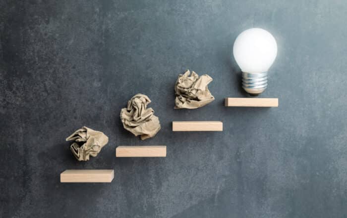 Conceptual image of crumpled paper balls as steps leading to a light bulb, symbolizing failure as part of the creative or problem-solving journey toward success