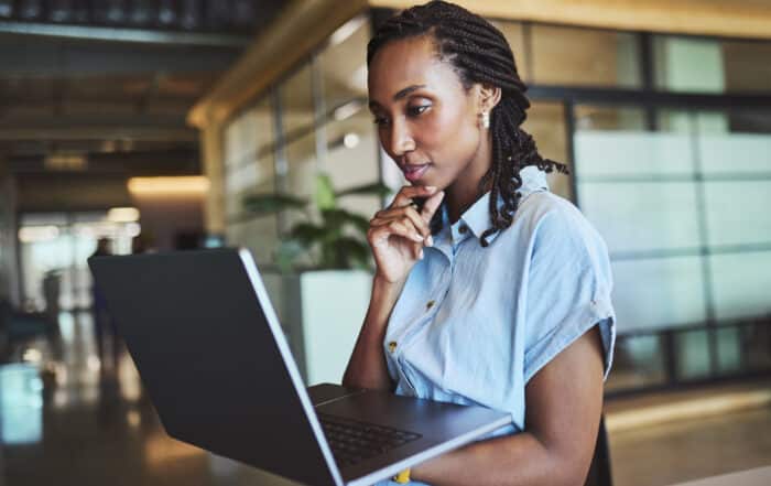 A thoughtful and focused young Black professional with braided hair stands while holding and looking down at her laptop, chin resting on her hand in a modern, open-plan office.